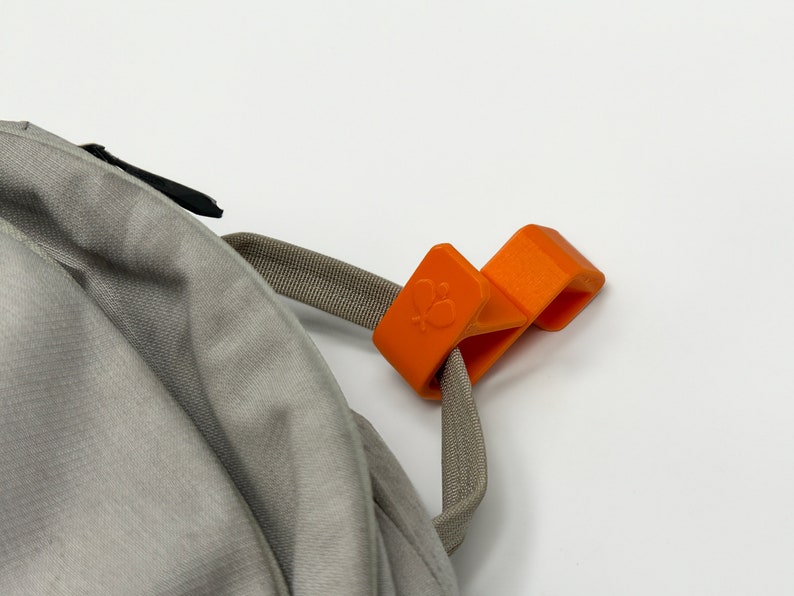 Puede incluir: Un clip naranja para mochila con un logotipo de mariposa est&aacute; sujeto a una correa gris y a una mochila gris. El clip est&aacute; hecho de un material pl&aacute;stico duro y tiene forma rectangular.