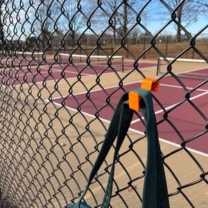 Puede incluir: Un soporte naranja para bolsas con clip, sujeto a una valla de alambre negra. Una bolsa verde oscuro cuelga del soporte. El fondo muestra una cancha de tenis con superficie roja y l&iacute;neas blancas, bajo un cielo despejado.