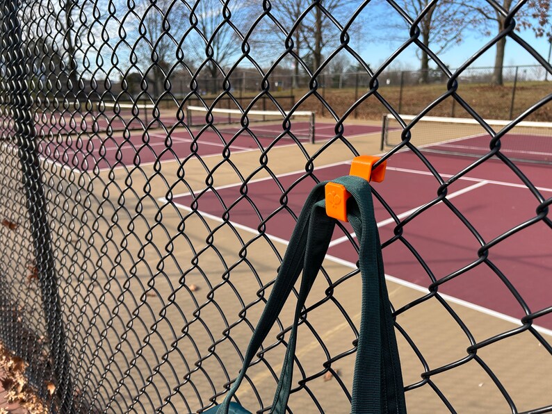 Puede incluir: Un soporte naranja para bolsas con clip, sujeto a una valla de alambre negra. Una bolsa verde oscuro cuelga del soporte. El fondo muestra una cancha de tenis con superficie roja y l&iacute;neas blancas, bajo un cielo despejado.