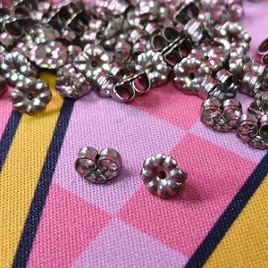 May include: A collection of silver-colored earring backs scattered on a pink and yellow patterned fabric. Two earring backs are in focus, showcasing their design. The earring backs have a flower-like shape with a central hole.