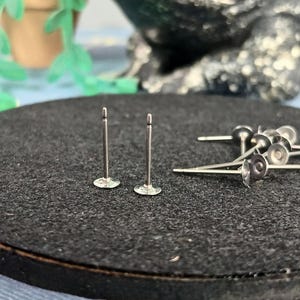 May include: Silver earring posts with flat, circular bases are displayed on a dark, textured surface. Two posts stand upright, with others scattered nearby. These small, delicate posts are suitable for jewellery making or crafting projects.