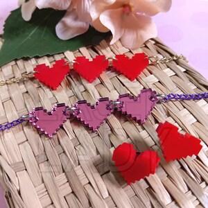 Puede incluir: Dos pulseras de coraz&oacute;n pixeladas en rojo y rosa, con cadenas doradas y plateadas, descansan sobre una superficie tejida. Tambi&eacute;n hay dos corazones pixelados rojos sueltos. Un accesorio encantador.