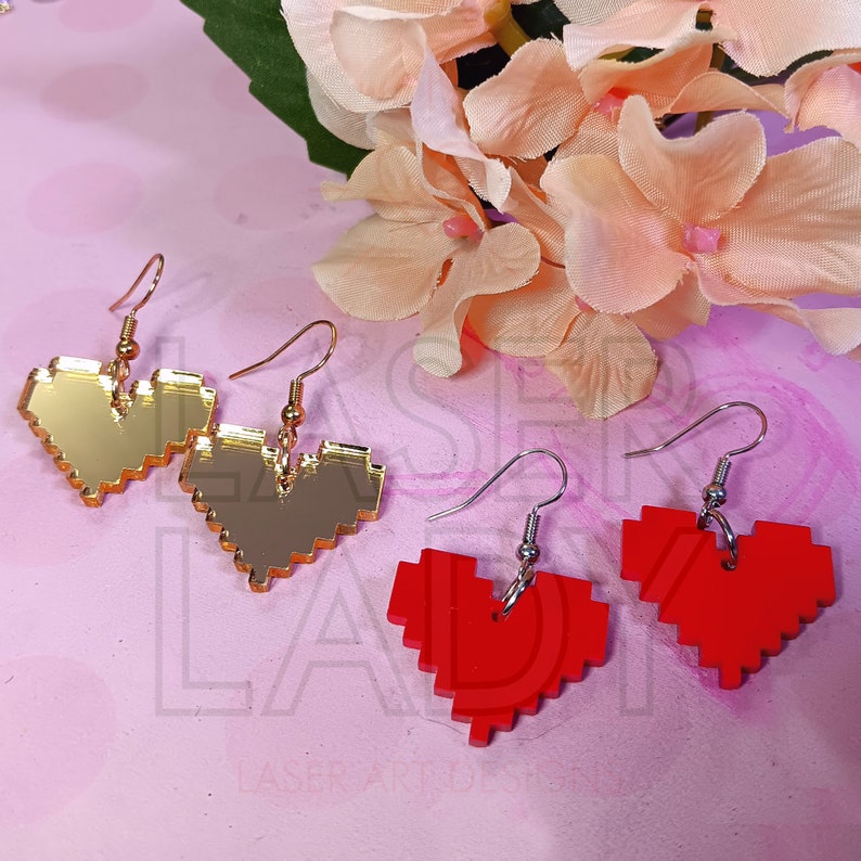 Puede incluir: Pendientes de coraz&oacute;n pixelados dorados y rojos. Los pendientes tienen una est&eacute;tica de videojuego retro. Los pendientes dorados tienen ganchos dorados, mientras que los pendientes rojos tienen ganchos plateados. Los pendientes se muestran sobre un fondo rosa con flores.