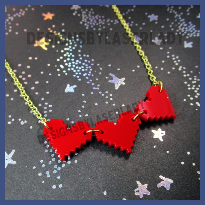 Puede incluir: Un collar de cadena dorada con tres colgantes rojos en forma de coraz&oacute;n. Los corazones tienen un dise&ntilde;o pixelado y est&aacute;n conectados por peque&ntilde;os anillos dorados. El collar se muestra sobre un fondo oscuro con acentos de estrellas. El texto en la imagen dice "DESIGNSBYLASERLADY".