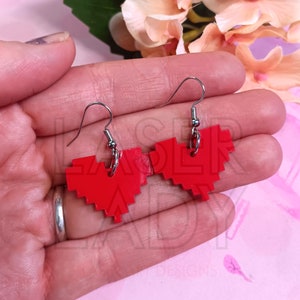 Puede incluir: Un par de pendientes rojos con forma de coraz&oacute;n pixelado con ganchos plateados. Los pendientes son de color rojo brillante y tienen una est&eacute;tica retro de videojuego. Los pendientes est&aacute;n sostenidos en una mano.
