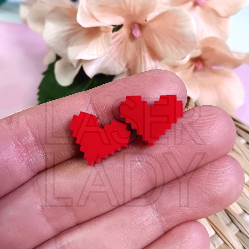 Puede incluir: Dos peque&ntilde;os objetos rojos en forma de coraz&oacute;n pixelados. Los corazones se muestran sobre una superficie de color claro, con un arreglo floral borroso en el fondo. Los corazones son de color rojo vibrante.