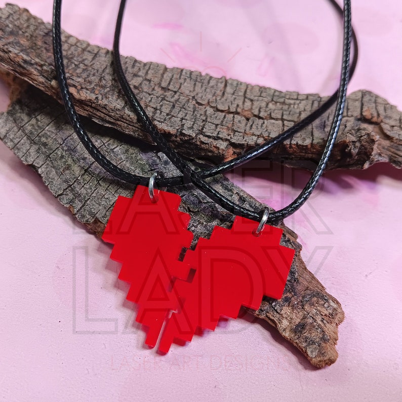 Puede incluir: Dos collares a juego con colgantes de coraz&oacute;n rojo pixelado y cordones negros. Los colgantes est&aacute;n hechos de un material rojo brillante y est&aacute;n dise&ntilde;ados para encajar. Los collares descansan sobre un trozo de madera sobre un fondo rosa.