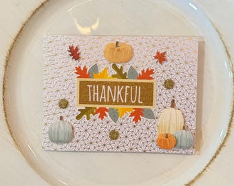 May include: A rectangular card with a geometric pattern and the word "THANKFUL" in gold glitter. The card is decorated with orange, yellow, and green leaves, pumpkins, and gold glitter accents. The card is placed on a white plate.