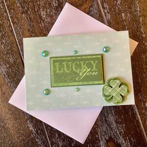 Könnte beinhalten: Eine St. Patrick's Day-Grußkarte mit den Worten "Lucky You" in Grün. Die Karte hat einen hellblauen Hintergrund mit Flamingomustern, ein grünes Kleeblatt und schillernde Edelsteine. Ein rosa Umschlag liegt darunter.