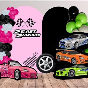 May include: A pink convertible car with a graphic design, a black tire, and several toy cars. The background includes balloons in black, pink, and green, and the text "2 Fast 2 Curious" with checkered flags.