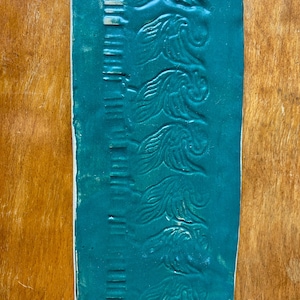 May include: A teal blue ceramic tile with a repeating pattern of stylised fish along the edge. The tile has a textured surface.