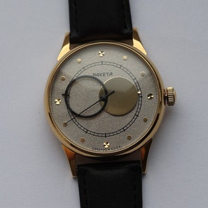 Puede incluir: Un reloj de pulsera dorado con una esfera blanca y agujas negras. El reloj tiene un diseño único con un círculo dentro de un círculo y la palabra "РАКЕТА" impresa en la esfera.