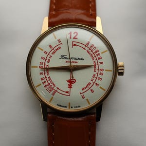 May include: A vintage gold-toned wristwatch with a cream-colored face and brown leather strap. The watch face features red markings and the word "Baltika". The watch is displayed on a white pillow.