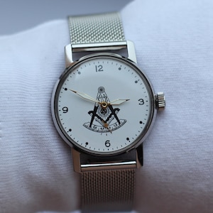 May include: A silver watch with a white face and a silver mesh band. The face of the watch features a black and white image of a compass and square, a symbol of Freemasonry.