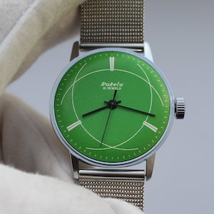 Puede incluir: Un reloj de pulsera plateado con una esfera verde y una correa de malla metálica. La esfera tiene marcas blancas y el texto "Raketa 21 JEWELS".