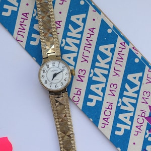 May include: A gold-tone wristwatch with a white face and black Roman numeral hour markers. The watch has a gold-tone metal band with a textured pattern. The watch is on a blue and white box with the text "ЧАЙКА" in pink and blue.