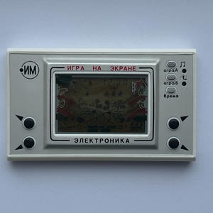Könnte beinhalten: Eine Vintage-Handheld-Elektronik-Spielkonsole mit einem grauen Kunststoffgehäuse. Der Bildschirm zeigt eine verpixelte Spielszene. Das Gerät hat schwarze Knöpfe und die Texte "ИГРА НА ЭКРАНЕ" und "ЭЛЕКТРОНИКА" sind sichtbar.