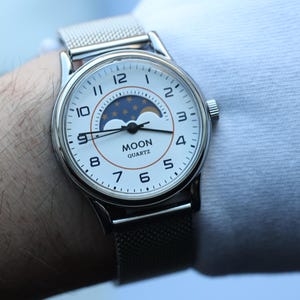 May include: A silver wristwatch with a white face featuring black numerals and hands. The watch has a mesh band and the words "MOON QUARTZ" are printed on the face. The watch face also displays a moon phase design.