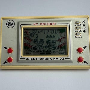Puede incluir: Un juego electrónico portátil vintage con un marco color crema y una pantalla que muestra una escena de dibujos animados. El juego tiene botones rojos y el texto "НУ, ПОГОДИ!" y "ЭЛЕКТРОНИКА ИМ 02". La hora mostrada es 12:00.
