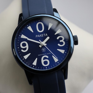 Puede incluir: Un reloj de pulsera azul marino con esfera redonda y correa a juego. La esfera presenta grandes números blancos en las posiciones de las 12, 3, 6 y 9, con la marca "PAKETA". El reloj está sobre una superficie blanca.