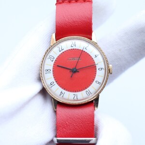 Peut inclure: Une montre-bracelet dorée avec un cadran rouge et des aiguilles noires. La montre a un anneau extérieur blanc avec des chiffres noirs et un bracelet en cuir rouge.