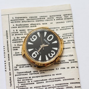 Puede incluir: Un reloj de pulsera dorado con una esfera negra y números blancos. La esfera del reloj dice "Rakieta" y tiene un segundero. El reloj está sobre un trozo de papel con texto en ruso.