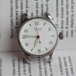 May include: A white analog wristwatch with a silver metal band. The watch face has black hour and minute hands, a red second hand, and green markings for the numbers. The watch face also has the text "Спортивные 17 камней" in Cyrillic script.