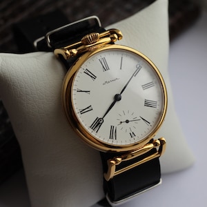 Può includere: Un orologio da polso dorato con quadrante bianco e indici orari romani neri. L'orologio ha un cinturino in pelle nera e dice "almann" sul quadrante.