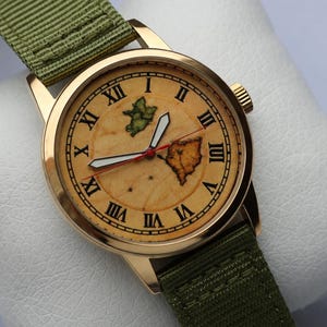 May include: A gold-toned wristwatch with a light brown face featuring two map illustrations and Roman numeral hour markers. The watch has white hands with a red second hand and a green fabric band.