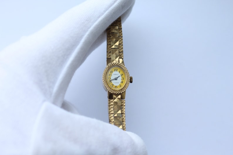 May include: A gold-tone wristwatch with a yellow face and black hands. The watch has a gold-tone metal band with a textured design.