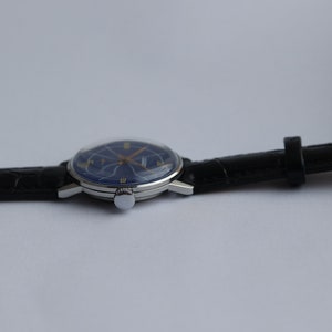 Puede incluir: Un reloj de pulsera plateado con una esfera azul y agujas doradas. El reloj tiene una correa de cuero negra.