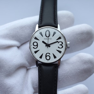 May include: A silver wristwatch with a white face and black numbers. The watch has a black leather strap and says "PAKEta" on the face.
