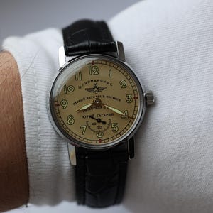 May include: A vintage wristwatch with a cream-colored face and black leather band. The watch face features black numerals, a second hand, and text in a foreign language. The watch is on a wrist with a white sleeve.