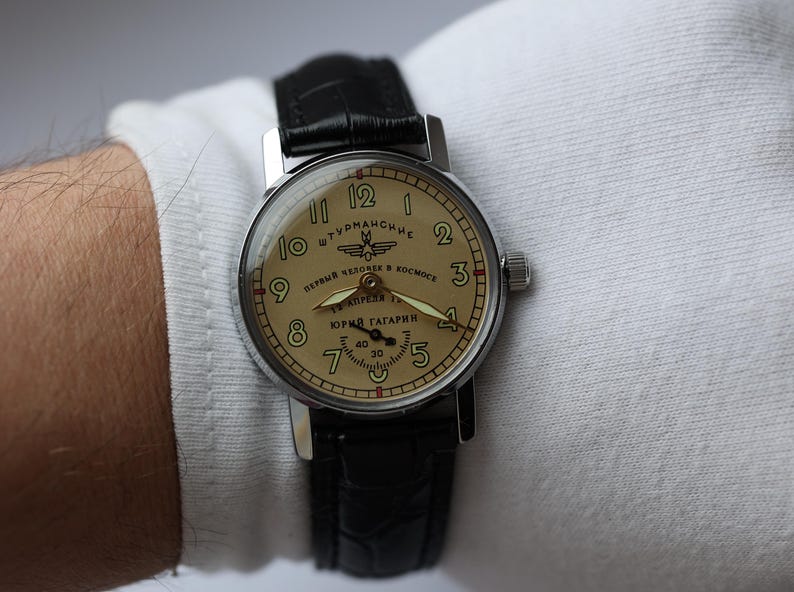 May include: A vintage wristwatch with a cream-colored face and black leather band. The watch face features black numerals, a second hand, and text in a foreign language. The watch is on a wrist with a white sleeve.