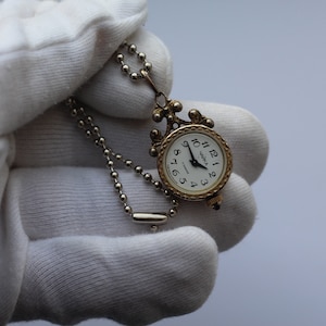 Puede incluir: Un reloj de bolsillo dorado con una esfera blanca y agujas negras. El reloj está en una cadena plateada y tiene un diseño decorativo alrededor del borde. El reloj está siendo sostenido en una mano enguantada de blanco.