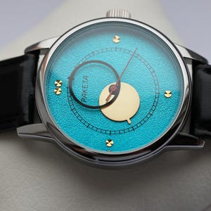 May include: A silver wristwatch with a black leather band. The watch face is turquoise blue with gold accents and a gold-colored moon phase indicator. The watch face also has the word "PAKETA" printed on it.