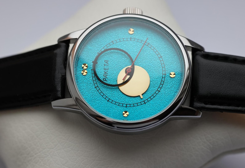 May include: A silver wristwatch with a black leather band. The watch face is turquoise blue with gold accents and a gold-colored moon phase indicator. The watch face also has the word "PAKETA" printed on it.