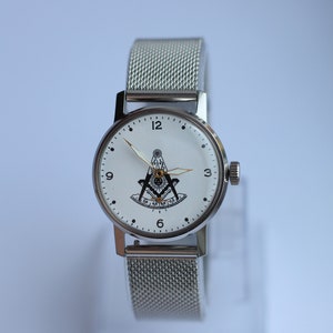 May include: A silver wristwatch with a white face and a silver mesh band. The face of the watch features a black and white compass and square design.