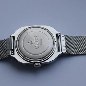 May include: Close-up of a silver-toned wristwatch with a mesh band. The watch back features the letters "CCCP" and a star emblem. The watch has a square face and a small crown.