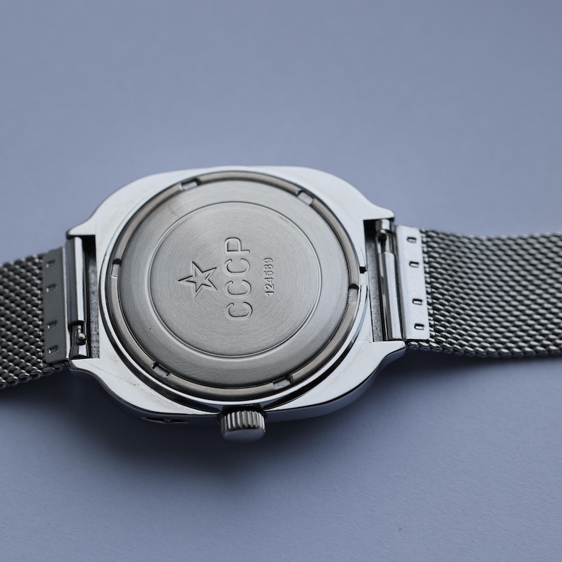 May include: Close-up of a silver-toned wristwatch with a mesh band. The watch back features the letters "CCCP" and a star emblem. The watch has a square face and a small crown.