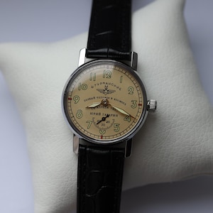 May include: A vintage wristwatch with a cream-colored face, black numerals, and a black leather band. The watch face features Cyrillic text and a small seconds dial. The watch is set on a white pillow.