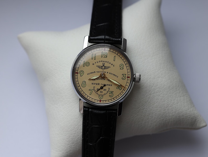 May include: A vintage wristwatch with a cream-colored face, black numerals, and a black leather band. The watch face features Cyrillic text and a small seconds dial. The watch is set on a white pillow.