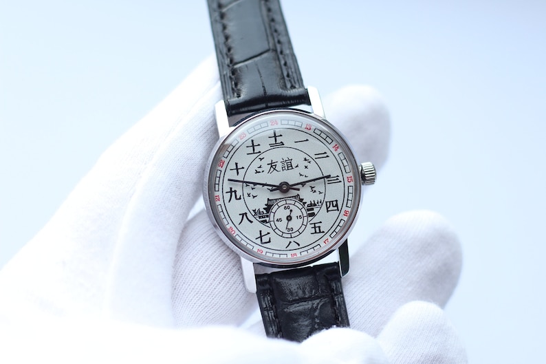 May include: A silver wristwatch with a white face and black leather band. The face features Chinese characters and a depiction of a building with birds flying around it.