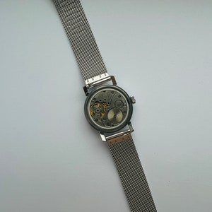 May include: A silver watch with a mesh band, showing the inner workings of the watch face.