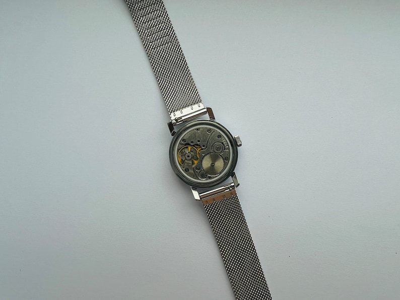 May include: A silver watch with a mesh band, showing the inner workings of the watch face.
