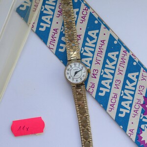 May include: A gold-tone wristwatch with a white face and black Roman numeral markings. The watch has a gold-tone metal band with a textured pattern. The watch is on a blue and white box with the text "Часы из Углича ЧАЙКА" in Russian.
