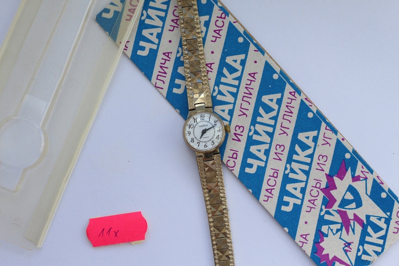 May include: A gold-tone wristwatch with a white face and black Roman numeral markings. The watch has a gold-tone metal band with a textured pattern. The watch is on a blue and white box with the text "Часы из Углича ЧАЙКА" in Russian.