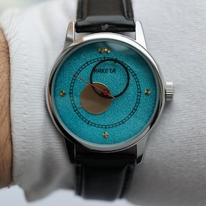 May include: A silver wristwatch with a blue face and black leather band. The watch face has a gold circle in the center and the word "Raketa" printed on it.