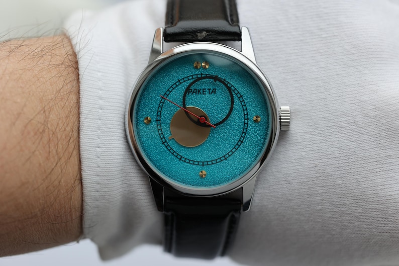 May include: A silver wristwatch with a blue face and black leather band. The watch face has a gold circle in the center and the word "Raketa" printed on it.