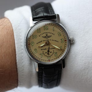 May include: A vintage wristwatch with a silver case and a tan face. The watch features black numerals, hands, and a black leather band. The watch face has Cyrillic text and the name "Yuri Gagarin".